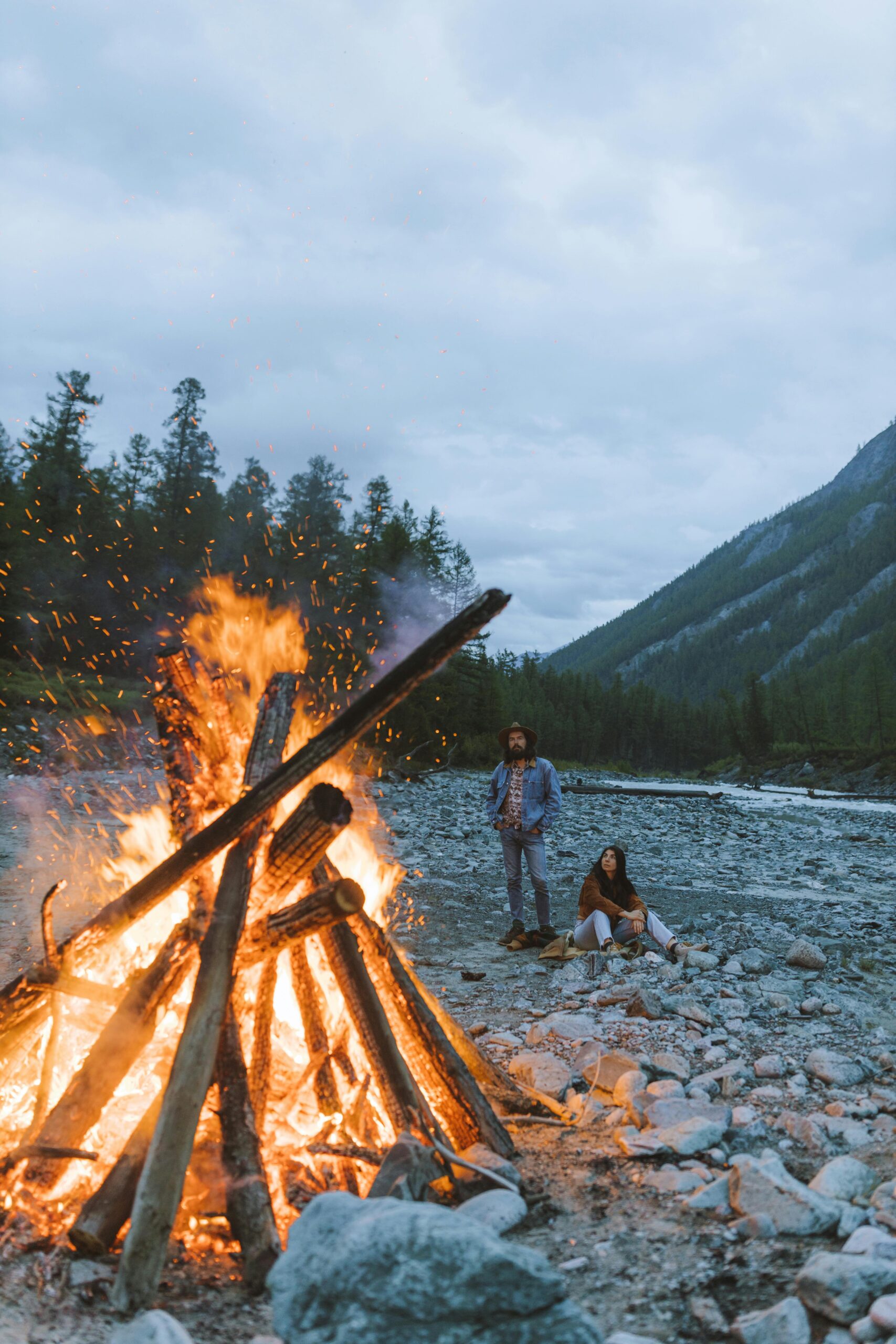 A couple sits by a large bonfire on a rocky riverside with mountains in the background.